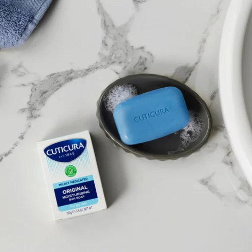 Cuticura Mildly Medicated Bar Soap on a bathroom sink with the packaging next to the blue bar of soap.